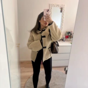 Beige teddyjacka med svarta detaljer - Storlek XS men passar större också. Beige teddyjacka med svarta kantdetaljer och knäppning framtill. Jackan har lång ärm och en oversized passform, perfekt för att hålla sig varm och snygg under kyliga dagar. Enkel att styla med både jeans och leggings.