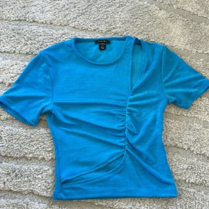 Blå t-shirt med rynkade detaljer - Säljer en blå t-shirt med korta ärmar och unik rynkad detalj framtill. T-shirten har en tight passform och är tillverkad i ett mjukt material som känns skönt mot huden. Perfekt för dig som vill sticka ut lite extra!a