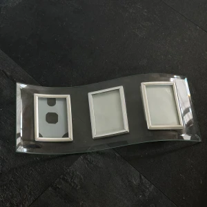 Böjd fotoram i glas med tre silverfärgade ramar - Modern fotoram i böjt glas med tre inbyggda rektangulära ramar i silverfärg. Perfekt för att visa tre foton tillsammans på ett stilrent sätt. Passar snyggt på hyllan eller skrivbordet. En bit av ena ramen har lossnat men det kan enkelt limmas ihop. 