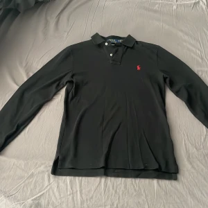 Svart pikétröja från Polo Ralph Lauren - Säljer en svart långärmad pikétröja från Polo Ralph Lauren i modellen Custom Slim Fit. Tröjan har klassisk krage, knappar vid halsen och röd broderad logga på bröstet. Perfekt för en stilren look.