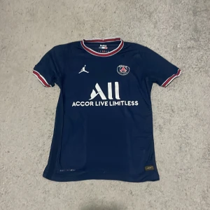 PSG matchtröja från Jordan - Mörkblå PSG fotbollströja med vita och röda detaljer, officiell logga på bröstet och Jordan-logga. Kortärmad modell med rund hals och tryckt sponsor på framsidan. Perfekt för dig som älskar fotboll och vill bära ditt lag.