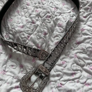 Silvrigt bälte med strass och mönstrat spänne - Säljer ett riktigt snyggt bälte i silver med massor av glittrande strass och detaljer längs hela bältet. Spännet är dekorerat med blommönster och stenar, perfekt för att ge outfiten extra bling.