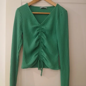 Grön ribbad topp med knyt från FB Sister - Säljer en grön långärmad topp från FB Sister. Toppen är ribbad med v-ringning och har snygg knyt- och rynkdetalj framtill som ger en cool look. Perfekt att styla med jeans eller kjol!