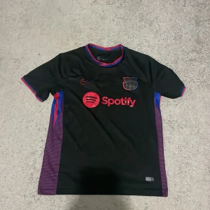 FC Barcelona svart fotbollströja Nike och den är custom-made - Säljer en svart FC Barcelona fotbollströja från Nike med röda och blå detaljer. Tröjan har korta ärmar, klubbmärke på bröstet och Spotify som sponsor. Snygga lila ränder på sidorna och färgade detaljer vid kragen.