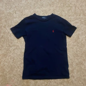 Mörkblå t-shirt från Polo Ralph Lauren - Mörkblå t-shirt från Polo Ralph Lauren med klassisk röd broderad logga på bröstet. Enkel och stilren design med rund hals och korta ärmar. Perfekt för en clean och tidlös look. Skriv till mig om ni har nån fråga eller nått om tröjan tröjan  eller om storlek för jag vet inte exakt vad storleken på tröjan är!