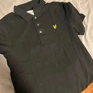 Svart pikétröja från Lyle & Scott - Säljer en klassisk svart pikétröja från Lyle & Scott med gul broderad logga på bröstet. Tröjan har krage, tre knappar och är kortärmad. Perfekt för en stilren och avslappnad look.