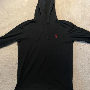 Svart hoodie från Polo Ralph Lauren - Svart hoodie från Polo Ralph Lauren med röd broderad logga på bröstet. Tröjan har långa ärmar, huva med dragsko och är tillverkad i mjukt material. Perfekt för en avslappnad stil. Passar xs/S