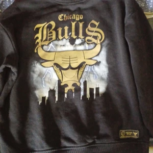 Svart Chicago Bulls sweatshirt från NBA - Cool svart sweatshirt med stort Chicago Bulls-tryck i guld och vitt på ryggen. Tröjan har rund halsringning och ribbade muddar. Motiv med blixtar, stadssilhuett och Bulls-logga. Perfekt för basketfans men även för en cool stil.