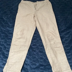 Beiga linnebyxor med snörning - Säljer ett par beiga linnebyxor med avslappnad passform och snörning i midjan. Perfekta för varma dagar och ger en skön, luftig känsla. Byxorna har raka ben och enkel design. Små i storleken 