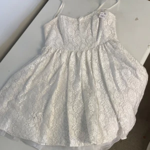 Vit spetsklänning från Paris Kate & I - Söt vit kortklänning från Paris Kate & I med smala axelband och vacker spets över hela klänningen. Klänningen har en dekorativ blomdetalj framtill och en feminin, lätt utställd passform. Perfekt för somriga tillfällen midsommar eller student.