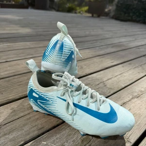 Blåa Nike fotbollsskor - Säljer ett par fotbollsskor från Nike med blåa detaljer och snörning. Skorna har en låg profil, rund tå och platt sula med dobbar för bra grepp på planen. Perfekta för dig som vill sticka ut på fotbollsplanen! Använda få tillfällen