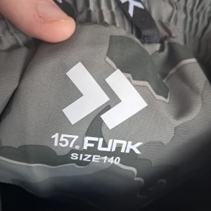 Grå camo täckbyxor från 157 Funk - Säljer ett par grå täckbyxor från 157 Funk i camouflagemönster. Byxorna har resår i midjan och är perfekta för utomhusaktiviteter under kallare dagar. Snygg och praktisk design för dig som gillar att vara ute.