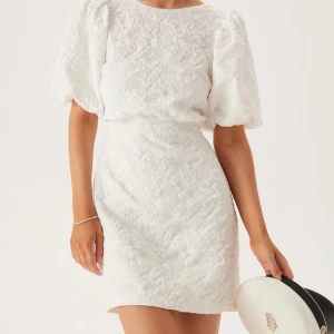 Vit kort klänning med puffärmar och rosett i ryggen - Superfin vit kort klänning med struktur, puffiga kortärmar och stor rosett i ryggen. Klänningen har öppen rygg och en figurnära passform. Perfekt för sommarens tillfällen och student. Klänningen är helt ny med prislapp då den var tänkt till skolavslutningen men hittade en annan, köptes ny för 599 kr😊💐