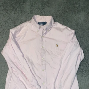 Rosa skjorta från Ralph Lauren - Snygg rosa skjorta från Ralph Lauren med klassisk krage och broderad logga på bröstet. Skjortan har långa ärmar och knappar framtill. Perfekt för en stilren look.