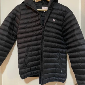 Svart pufferjacka från Guess - Snygg svart pufferjacka från Guess med dragkedja och huva. Jackan har en quiltad design och två praktiska fickor med dragkedja framtill. Perfekt för kyligare dagar.