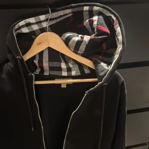 Svart hoodie från Burberry - Säljer en stilren svart hoodie från Burberry med klassiskt rutigt mönster i huvan. Den har en dragkedja framtill och är långärmad. Perfekt för en casual look. Jag har tyvärr växt ut denna. Pris kan diskuteras