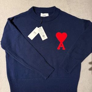 Mörkblå stickad tröja från Ami - Säljer en snygg mörkblå stickad tröja från Ami med ett rött hjärta och bokstaven A på bröstet. Tröjan har långa ärmar och en ribbad krage samt muddar. Perfekt för en stilren look.