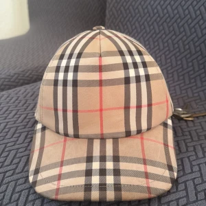 Rutig keps från Burberry äkta - Snygg keps från Burberry med klassiskt rutigt mönster i beige, svart och rött. Kepsen har en justerbar rem baktill för perfekt passform. Perfekt för att ge din outfit en stilren touch.
