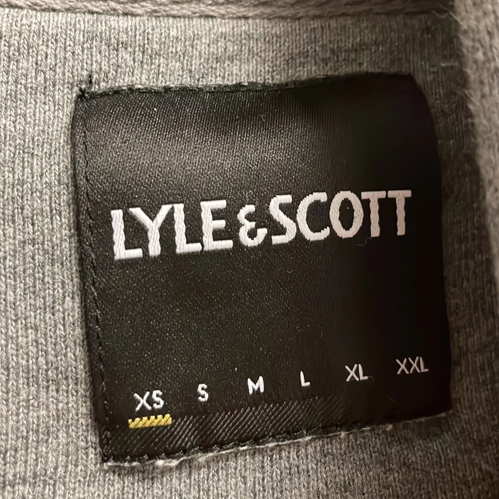 Säljer en stilren grå hoodie från Lyle & Scott med deras ikoniska gula logga på bröstet. Tröjan har en klassisk känguruficka och justerbar huva med dragsko. Perfekt för en avslappnad look.. Neuletakit & Villapaidat.