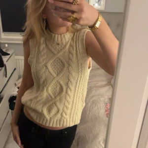 Beige stickad ärmlös topp - Säljer en snygg beige stickad topp med flätmönster. Den är ärmlös och har en ribbad kant vid halsen och nederdelen. Perfekt för lager-på-lager-stil eller som den är. Passar både till jeans och kjol!