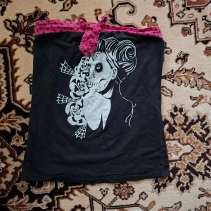 Svart tubtopp med tryck och rosa prickigt band - Cool svart tubtopp med ett unikt tryck av en dödskalle och blommor. Toppen har ett rosa band med svarta prickar som knyts i en rosett upptill. Perfekt för en edgy look!