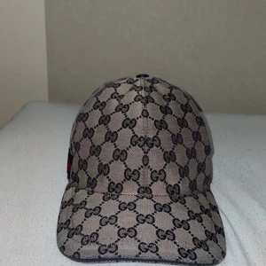 Grå beige keps från Gucci - Snygg grå beige keps från Gucci med det ikoniska GG-mönstret i svart. Kepsen har en klassisk design med en röd och blå randdetalj på sidan. Perfekt för att ge en lyxig touch till din outfit. PRIS GÅR DISKUTERA.