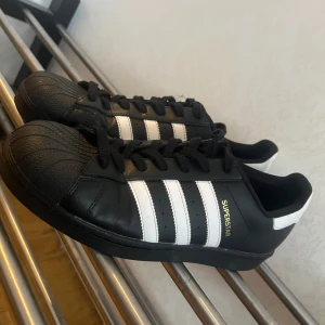 Svarta Adidas Superstar sneakers - Snygga svarta Adidas Superstar sneakers med de klassiska vita ränderna och guldfärgad text. Skorna har snörning och en ikonisk tåhätta. Perfekta för en stilig man. (PRISET KAN DISKUTERAS)