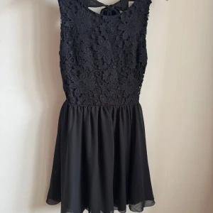 Svart spetsklänning från Model Behaviour - Snygg svart klänning med spetsdetaljer och öppen rygg från Model Behaviour. Klänningen är ärmlös och har en knytning i nacken. Perfekt för en elegant look.