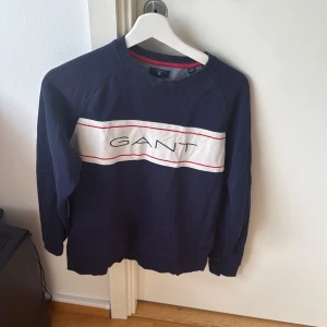 Mörkblå tröja från GANT - Säljer en snygg mörkblå tröja från GANT med vit panel och röd text på bröstet. Tröjan har långa ärmar och en klassisk passform. Perfekt för en stilren look.