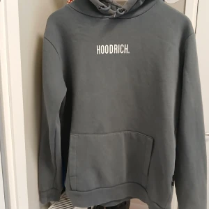 Grå hoodie från Hoodrich - Säljer en stilren grå hoodie från Hoodrich med vit text på bröstet. Den har en klassisk känguruficka och justerbar huva med dragsko. Perfekt för en avslappnad stil.