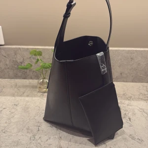 Svart axelväska - Snygg svart axelväska i skinn med en praktisk dragkedja framtill. Väskan har en justerbar axelrem och en stilren design som passar perfekt för alla tillfällen. Den är rymlig och har en modern look.