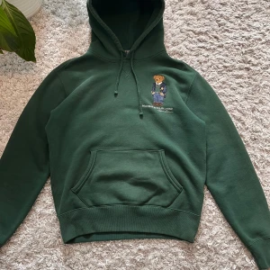 Grön hoodie från Polo Ralph Lauren - Säljer en snygg grön hoodie från Polo Ralph Lauren med en broderad nalle på bröstet. Hoodien har en klassisk känguruficka och en bekväm huva. Perfekt för en avslappnad stil. Passar perfekt för kyligare dagar.