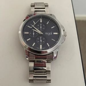 Silverfärgad klocka från Regal - Stilren klocka från Regal med silverfärgad metallarmband och mörk urtavla. Klockan har tre mindre urtavlor och tydliga visare. Perfekt för en elegant look. Sekundvisare är sönder men ingenting man tänker på, man kan fixa de själv eller gå till en klockmakare och fixa det billigt.