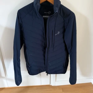 Mörkblå jacka från Pelle P - Säljer en mörkblå jacka från Pelle P med dragkedja och långärmade ärmar. Jackan har en quiltad design och en liten logotyp på bröstet samt en på ärmen. Perfekt för kyligare dagar.
