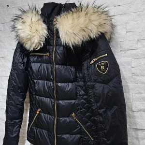 Rock and blue jacka NY - Snygg svart dunjacka med fluffig päls på huvan och guldfärgade dragkedjor. Jackan har en quiltad design och flera praktiska fickor. Perfekt för kyliga dagar när du vill hålla stilen.