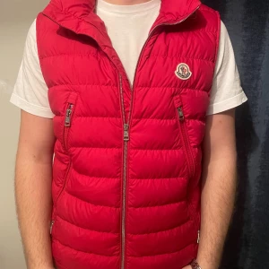 Röd väst från Moncler - Säljer en snygg röd dunväst från Moncler med dragkedja och logotyp på bröstet. Perfekt för den kommande våren å sommarn. Västen har en quiltad design och praktiska fickor med dragkedja. Nypris 10 000kr