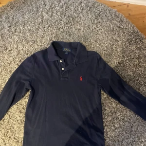 Mörkblå pikétröja från Ralph Lauren - Säljer en klassisk mörkblå pikétröja från Ralph Lauren med det ikoniska röda broderade logotypen på bröstet. Tröjan har långa ärmar och en stilren krage med knappar. Perfekt för en avslappnad men stilren look.