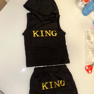 Svart ärmlös huvtröja med texten 'KING' - Snygg svart ärmlös huvtröja med texten 'KING' i gult på bröstet. Perfekt för en cool och avslappnad stil. Matchande svarta shorts ingår. Passar barn mellan 1 år och 3. 