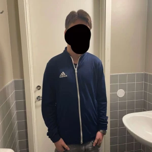 Blå träningsjacka från Adidas - Säljer en blå träningsjacka från Adidas med dragkedja och vit logga på bröstet. Jackan har långa ärmar och en stilren design, perfekt för sportiga aktiviteter. Den är tillverkad i ett syntetmaterial som ger en bekväm passform.
