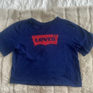 Blå t-shirt från Levi's - Säljer en blå t-shirt från Levi's med det klassiska röda logotyptrycket på framsidan. T-shirten är kortärmad och tillverkad i bomull, perfekt för en avslappnad stil. Passar bra till jeans eller shorts för en cool look. Den är också lite kortare än en vanlig tshirt!