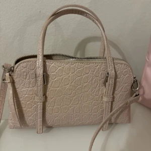 Beige axelväska med krokodilmönster - Snygg beige axelväska med krokodilmönster och dragkedja. Väskan har både handtag och en justerbar axelrem för flexibel användning. Perfekt för att bära dina nödvändigheter med stil.