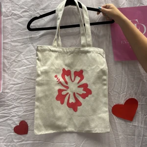 Beige shoppingväska med röd blomma - Vi är ett UF företag som säljer tygväskor med handmålade tryck på framsidan. Perfekt för att bära med sig det nödvändigaste med stil.