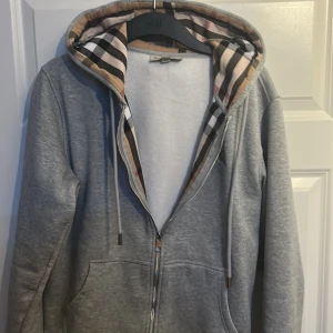 Grå huvtröja från Burberry - Säljer en stilren grå huvtröja från Burberry med dragkedja och klassiskt rutigt mönster i huvan. Tröjan har långa ärmar och ribbade muddar. Perfekt för en avslappnad look.