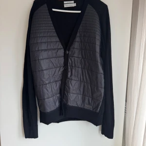 Svart och grå jacka/kofta från Peak Performance - Snygg svart och grå jacka från Peak Performance med quiltad framsida och knappar. Tillverkad i merinoull 