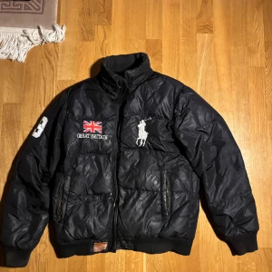Svart dunjacka från Polo Ralph Lauren - Säljer en svart dunjacka från Polo Ralph Lauren med broderad logga och Union Jack-flagga på bröstet. Jackan har dragkedja och ribbade muddar. Insidan är fodrad i beige. Perfekt för kyliga dagar! Löparen till dragkedjan fattas