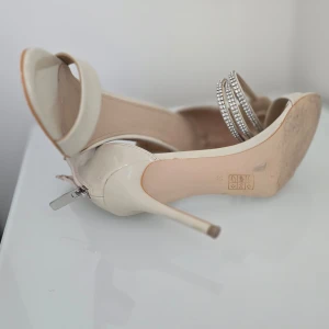 Beige klackskor från Guess - Eleganta beige klackskor från Guess med smal klack och öppen tå. Skorna har en glittrig rem runt ankeln och dragkedja bak för enkel påtagning. Perfekta för att ge en touch av glamour till din outfit.