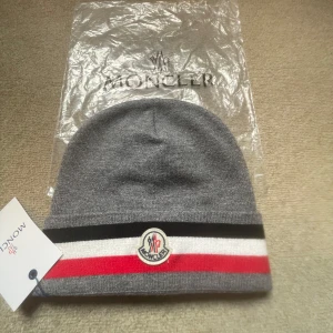 Grå mössa från Moncler - Snygg grå mössa från Moncler med röd, vit och svart randdetalj. Den har en broderad Moncler-logga framtill. Perfekt för att hålla sig varm med stil.oanvänd och. såklart e den ny såklart kan priset sänkas vid snabbt köp