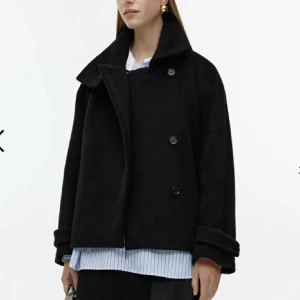 Svart kappa från Arket - Snygg svart kappa från Arket med dubbelknäppning och bred krage. Tillverkad i ull från Manteco, vilket ger en lyxig känsla. Perfekt för våren med sin stilrena design. Kan mötas upp eller skickas!