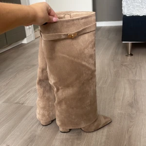Beige mockastövlar med klack - Snygga beige stövlar i mocka med hög klack och dragkedja på sidan. De har en dekorativ rem med gulddetalj upptill. Perfekta för att ge en elegant touch till din outfit. Aldrig använt skriv för fler bilder💕