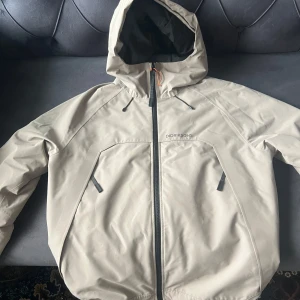 Beige regnjacka från Didriksons - Säljer en beige regnjacka från Didriksons med huva och dragkedja. Jackan är 100% vattentät och har en stilren design med två fickor framtill. Perfekt för regniga dagar! (Priset kan diskuteras).